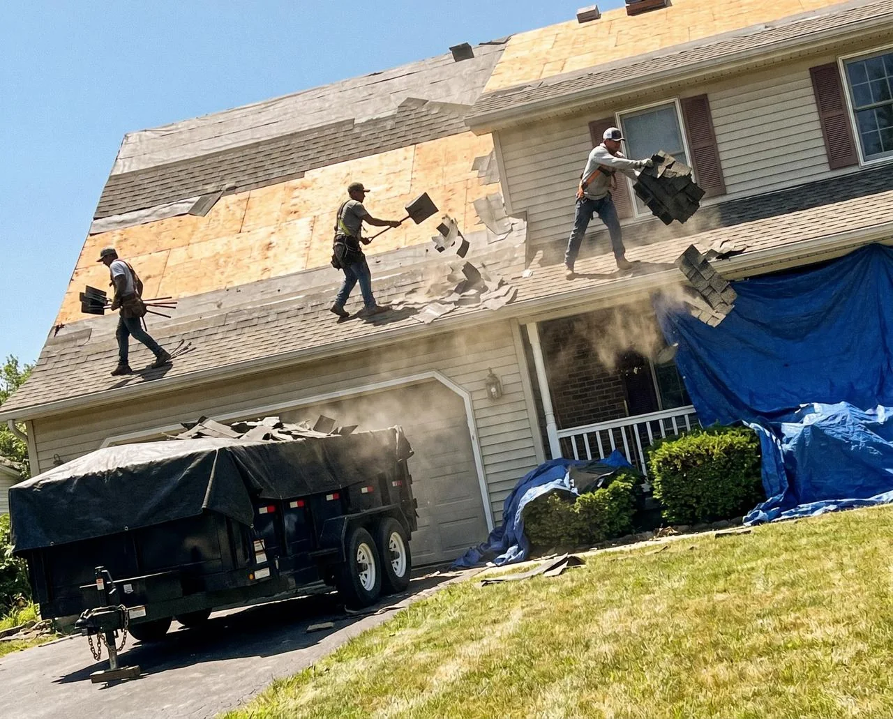 Workers removing old shingles from a house roof with a dump trailer in the driveway